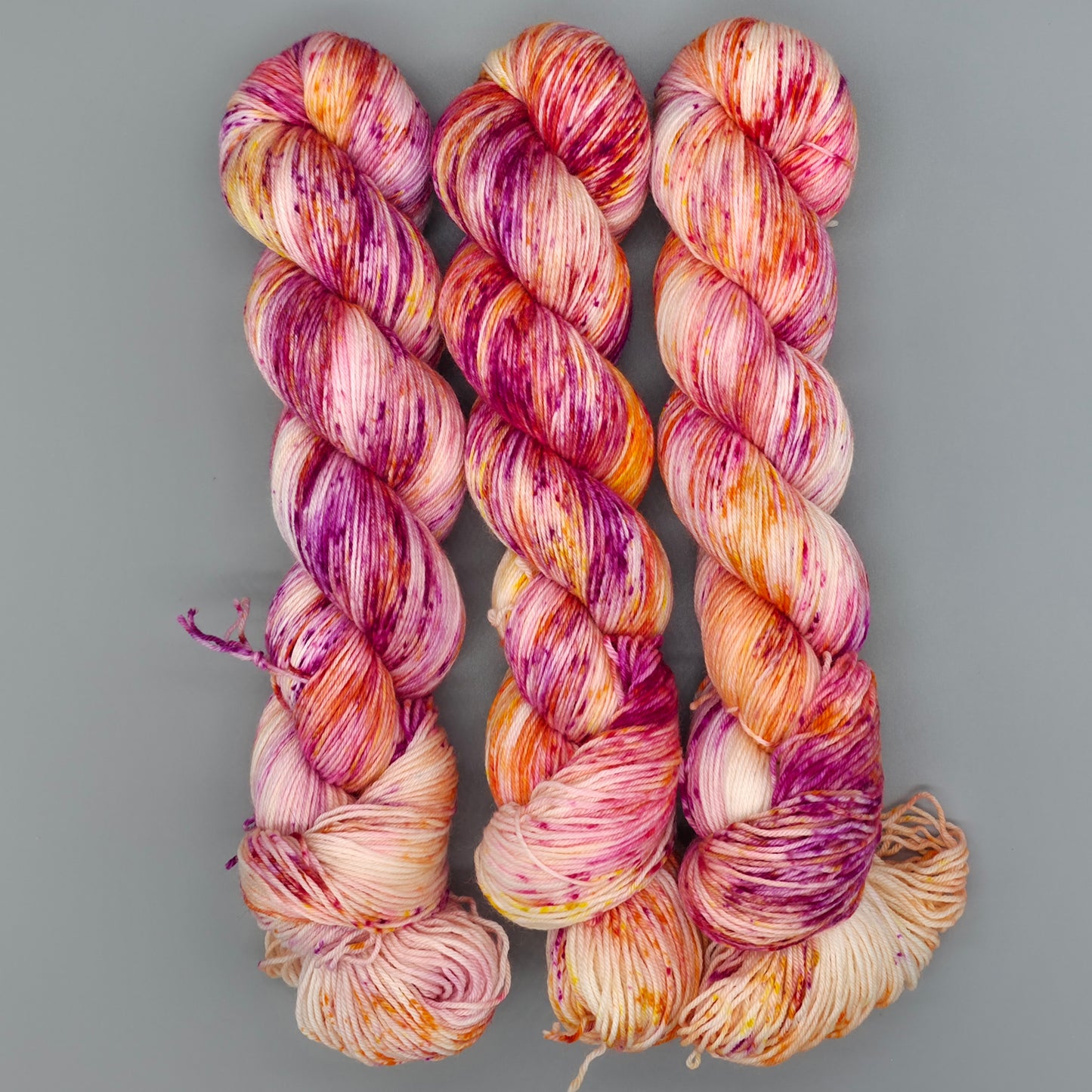 Three skeins of variegated yarn with pink, purple, orange, and cream tones, arranged vertically on a solid gray background.